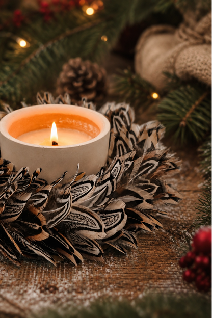 A lit white candle in a rustic feathered holder, surrounded by pinecones, evergreen branches, red berries, and warm string lights on a wooden surface.