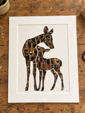 A mother deer and her fawn silhouette cutout, backed with natural real hen pheasant feathers, displayed on a rustic wooden workbench.