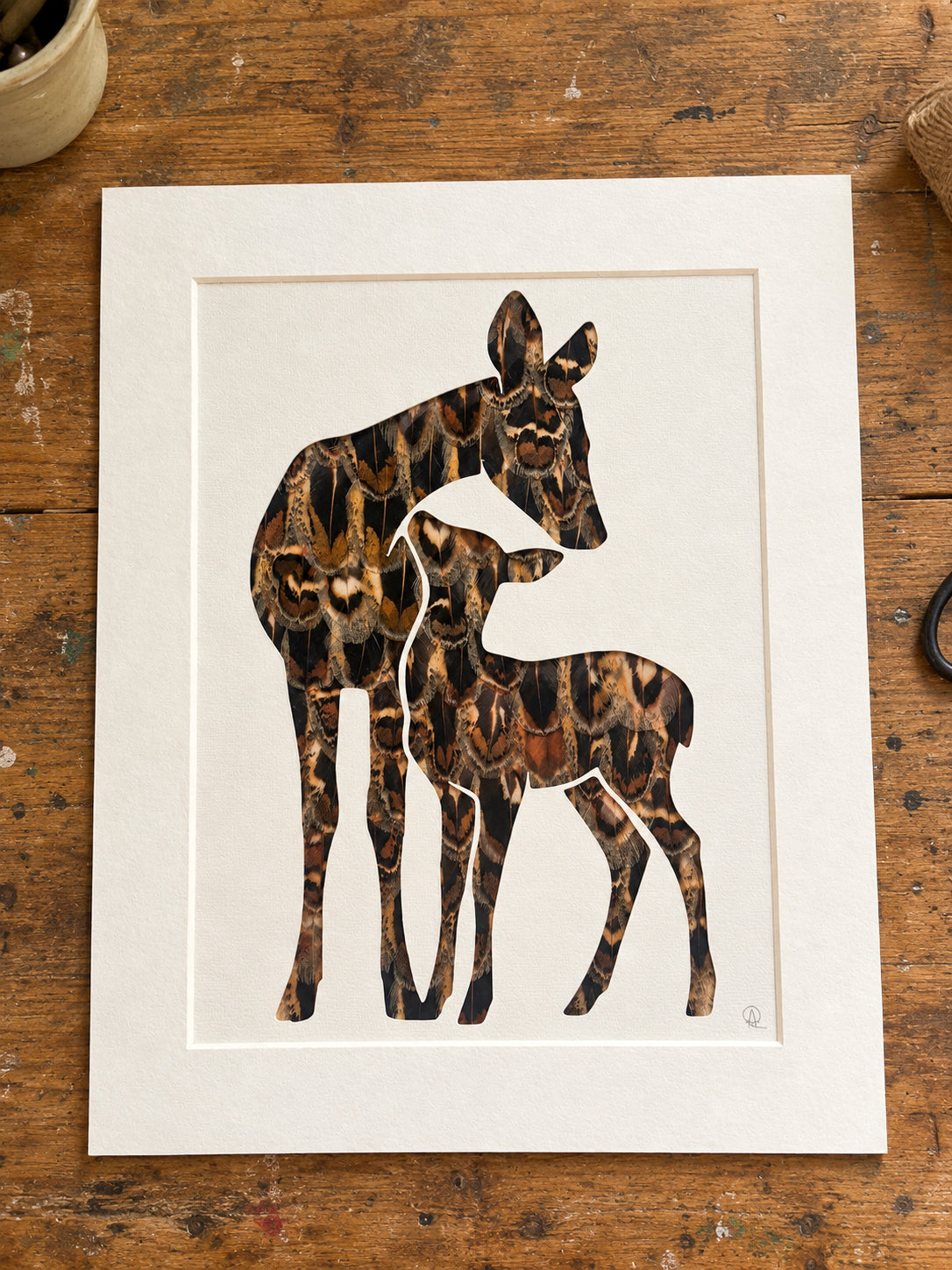 A mother deer and her fawn silhouette cutout, backed with natural real hen pheasant feathers, displayed on a rustic wooden workbench.
